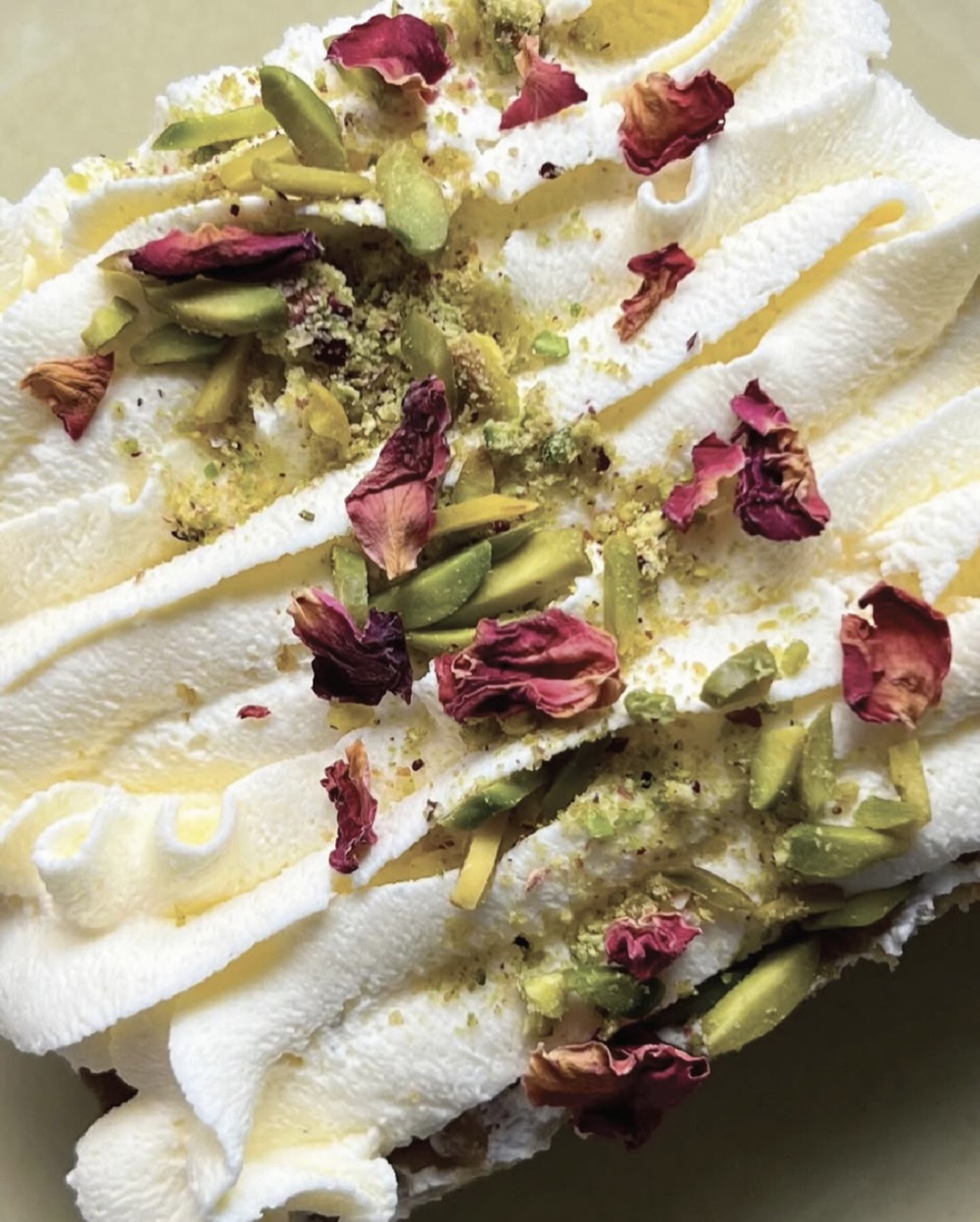 A slice of milk cake topped with whipped cream, slivered pistachios, and dried rose petals.
